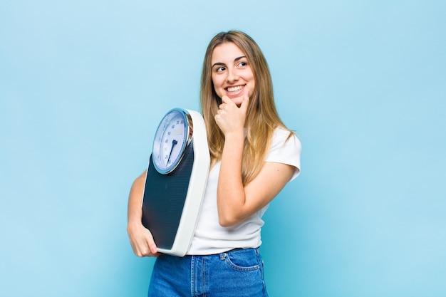 Blonde hübsche Frau, die mit einem glücklichen, selbstbewussten Ausdruck mit der Hand am Kinn lächelt, sich wundert und zur Seite schaut