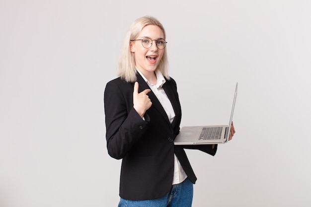 Blonde hübsche Frau, die sich glücklich fühlt und auf sich selbst zeigt, aufgeregt und hält einen Laptop