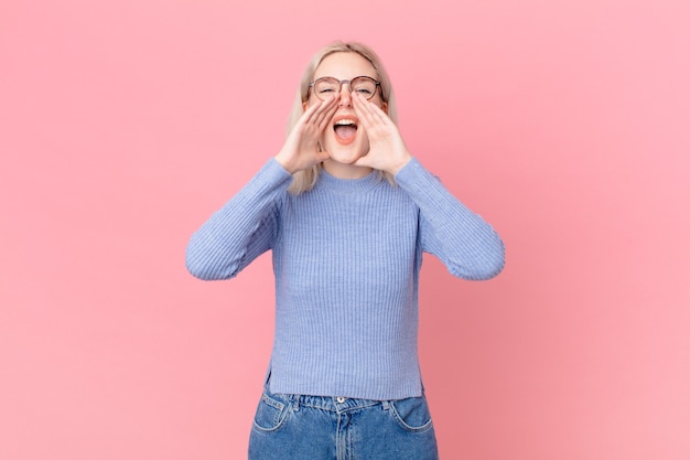 Blonde hübsche Frau, die sich glücklich fühlt und mit den Händen neben dem Mund einen großen Schrei ausstößt