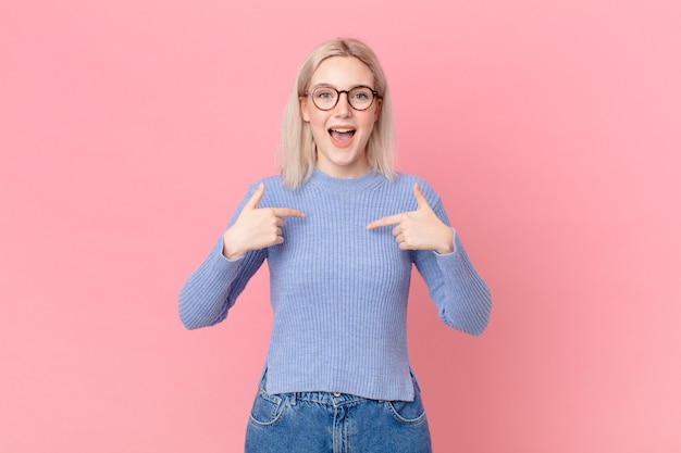 Blonde hübsche Frau, die sich glücklich fühlt und mit einem aufgeregten auf sich selbst zeigt