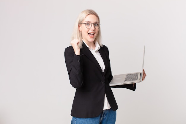 Blonde hübsche Frau, die sich schockiert fühlt, lacht und Erfolg feiert und einen Laptop hält