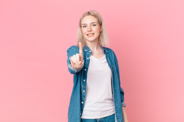 Blonde hübsche Frau, die stolz und selbstbewusst lächelt und die Nummer eins macht