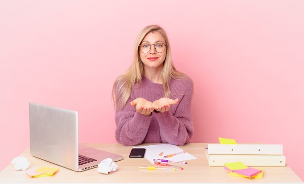 Blonde hübsche Frau junge blonde Frau, die glücklich mit freundlichem Lächeln lächelt und ein Konzept anbietet und zeigt und mit einem Laptop arbeitet