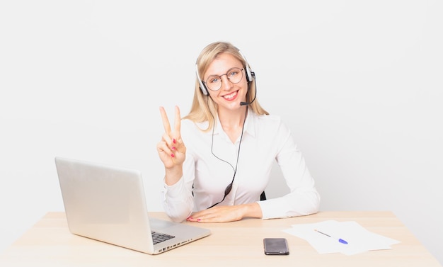 Blonde hübsche Frau junge blonde Frau, die lächelt und glücklich aussieht, Sieg oder Frieden gestikuliert und mit einem Laptop arbeitet