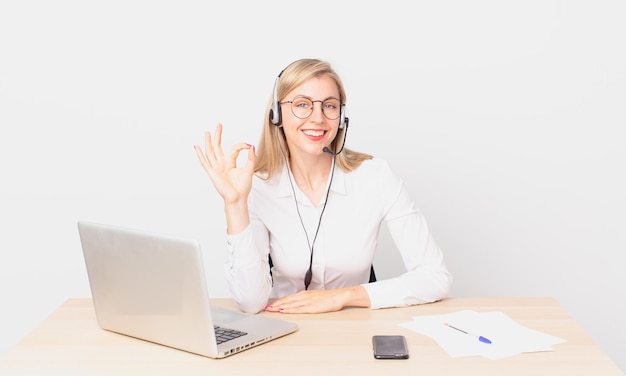 Blonde hübsche Frau junge blonde Frau, die sich glücklich fühlt, Zustimmung mit okayer Geste zeigt und mit einem Laptop arbeitet