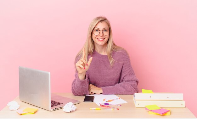 Blonde hübsche Frau junge blonde Frau, die stolz und selbstbewusst lächelt, die Nummer eins macht und mit einem Laptop arbeitet