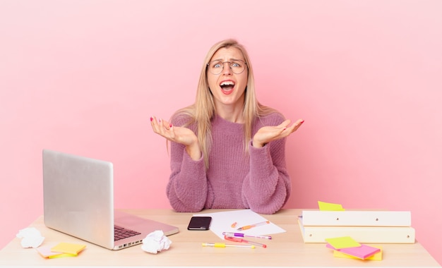 Blonde hübsche Frau junge blonde Frau, die verzweifelt, frustriert und gestresst aussieht und mit einem Laptop arbeitet