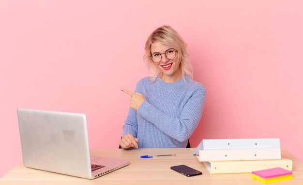 Blonde hübsche Frau junge hübsche Frau, die aufgeregt und überrascht aussieht und auf die Seite zeigt. Arbeitsplatz-Schreibtisch-Konzept