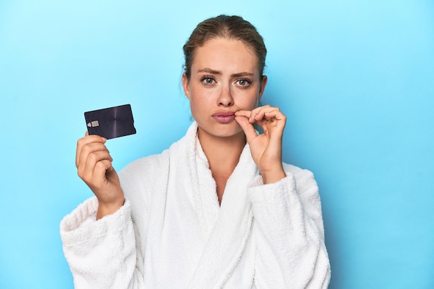 Blonde in Bademantel mit Kreditkarte in blauem Studio mit Fingern auf den Lippen, die ein Geheimnis bewahren