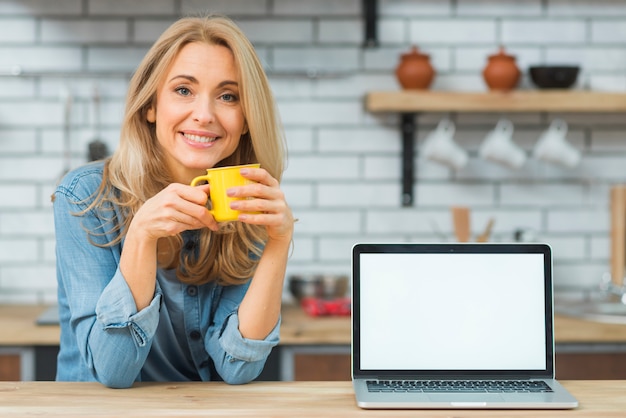 Foto blonde junge frau, die tasse kaffee mit laptop auf holztisch hält