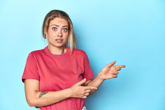 Blonde junge Frau in rotem T-Shirt, schockiert und mit dem Zeigefinger auf einen Kopierplatz zeigt