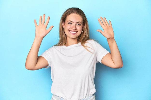 Blonde junge Frau in weißem T-Shirt auf blauem Hintergrund zeigt Nummer zehn mit Händen