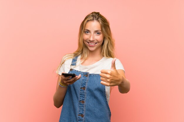 Blonde junge Frau mit einem Handy über lokalisierter rosa Wand mit den Daumen oben, weil etwas Gutes geschehen ist