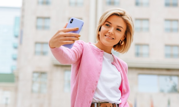 Blonde junge Frau trägt blaue Jeans, weißes T-Shirt und rosa Jacke, lächelt und macht Selfie auf dem Smartphone Fröhliche Frau, die im Frühling auf der Straße der Stadt Selbstporträt macht