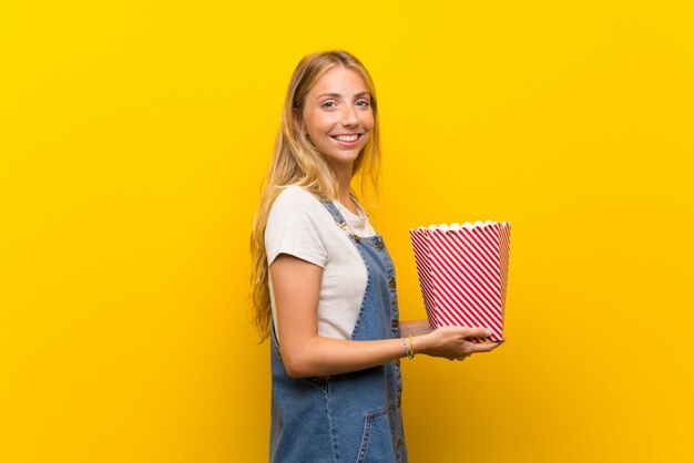 Blonde junge Frau über der lokalisierten gelben Wand, die eine Schüssel Popcorn hält