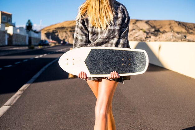 Blonde junge Frau von hinten gesehen, die in einer kleinen Stadt in der Nähe eines Berges mit einem Skateboard die Straße hinuntergeht?