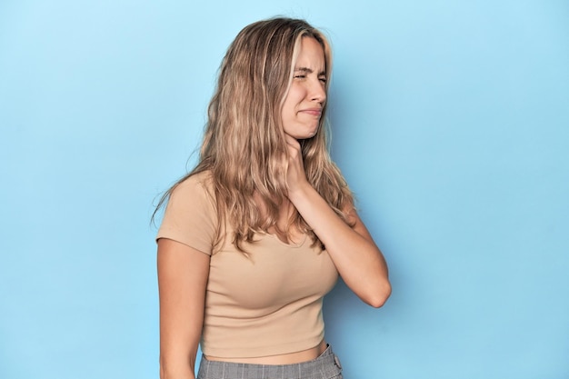 Blonde junge weiße Frau im blauen Studio leidet an Halsschmerzen aufgrund eines Virus oder einer Infektion