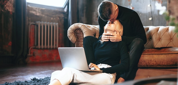 Blonde kaukasische frau, die auf dem boden mit einem computer arbeitet, während ihr geliebter sie küsst