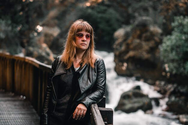 Foto blonde kaukasische junge frau mit sonnenbrille, schwarzer jacke und schwarzem pullover, die sich auf das holz lehnt