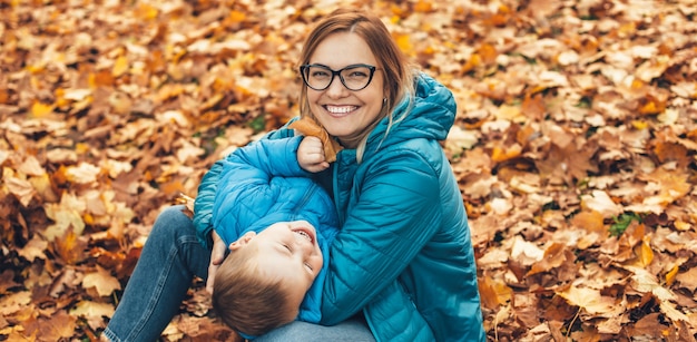 Blonde kaukasische Mutter mit Brille in blauen Kleidern und ihrem Sohn, der auf dem Boden mit Blättern spielt