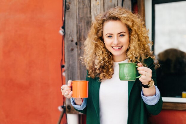 Blonde, lockige Frau mit Kaffeetassen, glücklichem Gesichtsausdruck und angenehmem Lächeln, bereit, Freunde zu verwöhnen