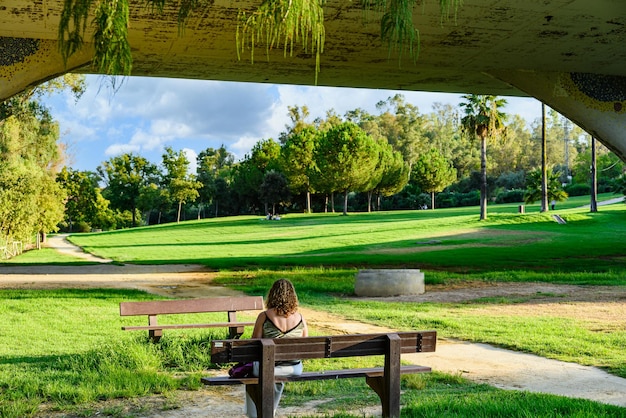 Blonde lockige Frau sitzt auf einer Bank im Park La Retama in Alcala de Guadaira, Sevilla, Spanien.