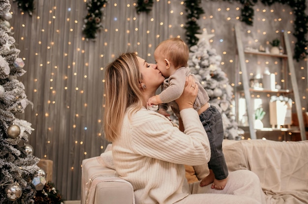 Blonde Mama im Strickanzug spielt mit einem kleinen Jungen auf dem Sofa in einem Zimmer mit Weihnachtsdeko