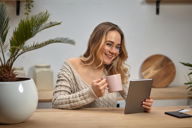 Blonde Model Bloggerin Brille beige Strickpullover Lächelnd Tablette rosa Tasse halten Sitzen in der Küche am Holztisch mit Palme im weißen Topf Smartphone und Plastikkarte drauf Nahaufnahme