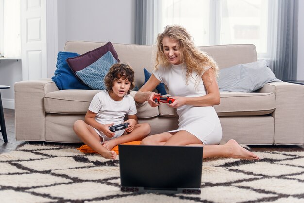 Blonde Mutter und ihr kleiner lustiger Sohn spielen Videospiele auf der Konsole. Glückliche Familienzeit zusammen.