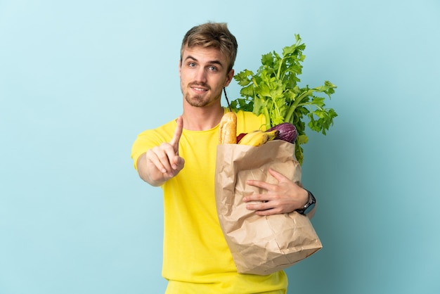 Blonde Person, die eine Tüte Essen zum Mitnehmen nimmt, das auf blauer Wand isoliert wird, die einen Finger zeigt und hebt