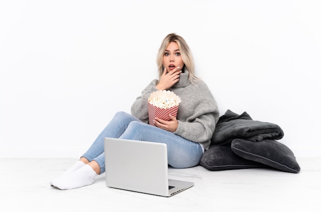 Blonde Teenagerfrau, die Popcorn isst, während sie einen Film auf dem Laptop sieht, überrascht und schockiert, während sie richtig schaut