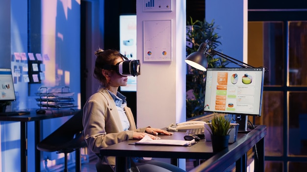 Blonde Unternehmerin analysiert Diagramme mithilfe eines VR-Headsets und arbeitet nach Feierabend im Geschäftsbüro. Junger Erwachsener mit Virtual-Reality-Brille plant Finanzbericht am Computer.