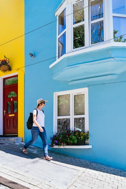 Blonde, wanderlustige, fröhliche Frau erkundet mit Sonnenbrille, Hut und Rucksack farbenfrohe Straßen inmitten hübscher blauer und gelber Häuser mit floralen Akzenten