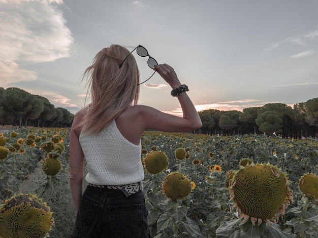 Blondehaired Frau im Sonnenblumenfeld bei Sonnenuntergang mit Sonnenbrille in ihren Händen