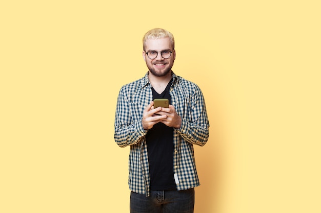 Blonder bärtiger Mann mit Brille lächelt in die Kamera, während er auf einem Handy plaudert, das an einer gelben Wand posiert