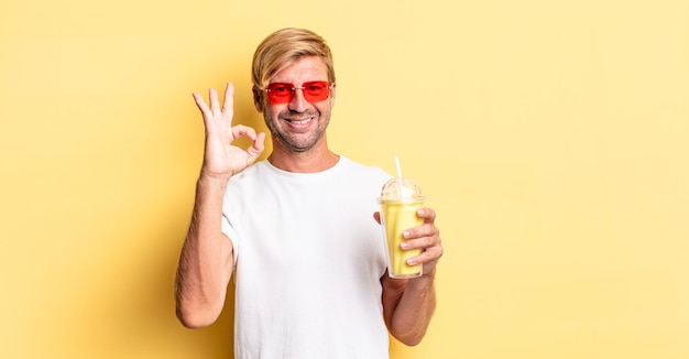 Blonder erwachsener Mann, der sich glücklich fühlt und Zustimmung mit einer okayen Geste mit einem Milchshake zeigt