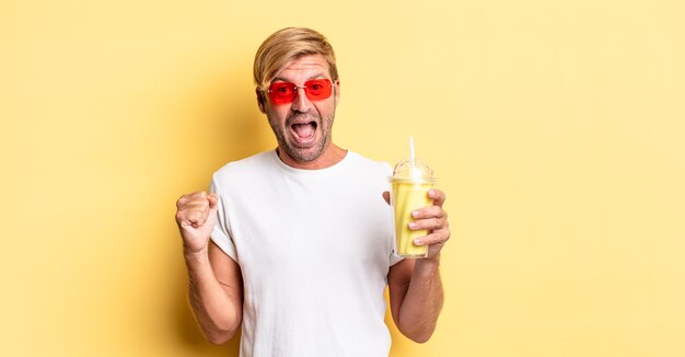 Blonder erwachsener Mann, der sich schockiert fühlt, lacht und den Erfolg mit einem Milchshake feiert