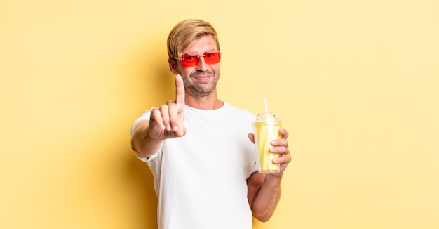 Blonder erwachsener Mann, der stolz und selbstbewusst lächelt und mit einem Milchshake die Nummer eins macht