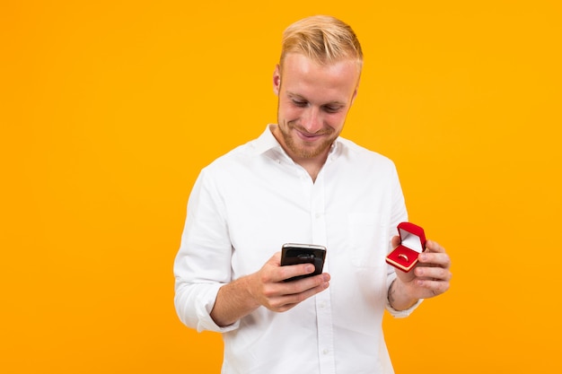 Blonder europäischer Mann macht ein Angebot, das einen Ring in einem Kasten und in einem Telefon auf einem Gelb hält.