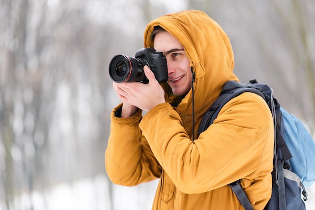 Blonder Fotojunge in der Kapuze, der Winterbäume mit Schnee schießt, während er im Wald spazieren geht