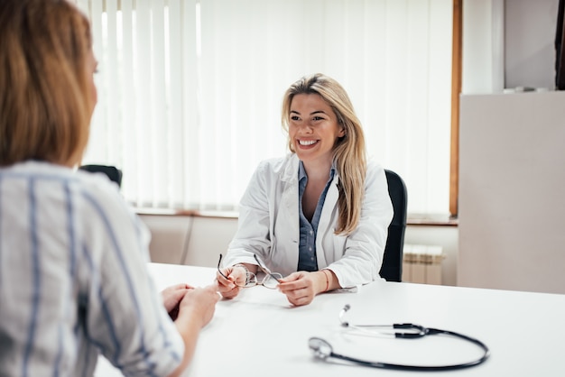 Blonder Hausarzt, der Brillen bei der Unterhaltung mit einer Patientin hält.