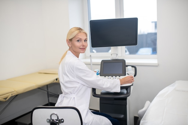 Blonder hübscher Ultraschallspezialist im weißen Gewand, der am Ultraschallgerät sitzt und auf den Patienten wartet