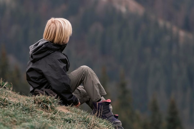 Blonder Junge auf Wanderung während der Pause Kind in Trekkingkleidung entspannt sich am Berghang Junge sitzt auf einem Hügel