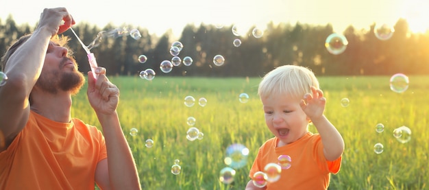 Blonder Junge des netten Kleinkindes und sein Vater, die mit Seifenblasen auf Sommerfeld spielt. Schönes Sonnenunterganglicht. Happy Kindheit Konzept. Authentisches Lifestyle-Image