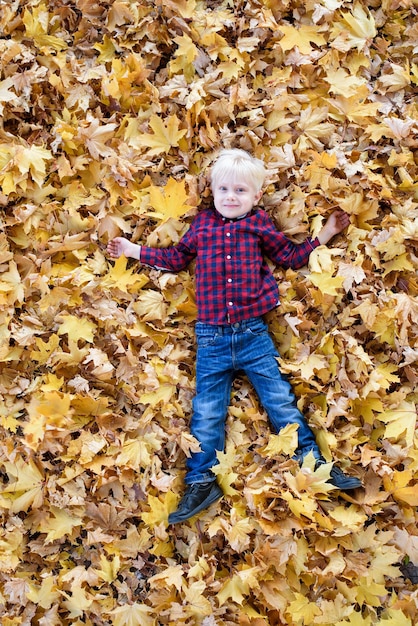 Blonder Junge in einem karierten Hemd liegt in gelben Herbstblättern Draufsicht Herbstkonzept