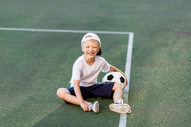 Blonder Junge in einer Mütze in einer Sportuniform sitzt auf einem Fußballfeld mit einem Fußball