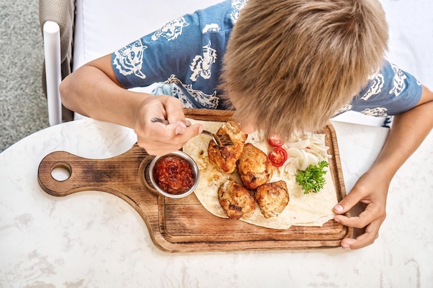 Blonder Junge isst gegrilltes Fleischgericht, das am Tisch im Restaurant in der Nähe der oberen Ansicht sitzt