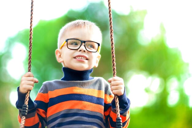 Blonder Junge mit Brille auf einer Schaukel im Park Die Freude