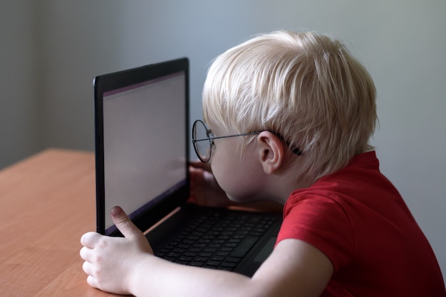 Blonder Junge mit Brille sitzt seine Nase in einem Laptop begraben. Internet und Vorschulkind