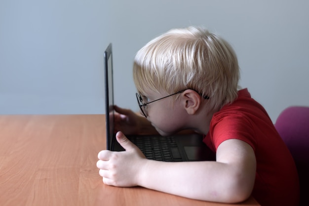 Blonder Junge mit Brille sitzt seine Nase in einem Laptop begraben. Internet und Vorschulkind